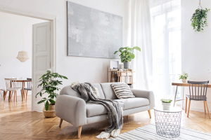 Bright and airy modern living room with a light grey sofa adorned with a cozy knitted throw blanket and striped cushion, a large abstract painting on the wall, light hardwood floors, and green plants adding a touch of nature.
