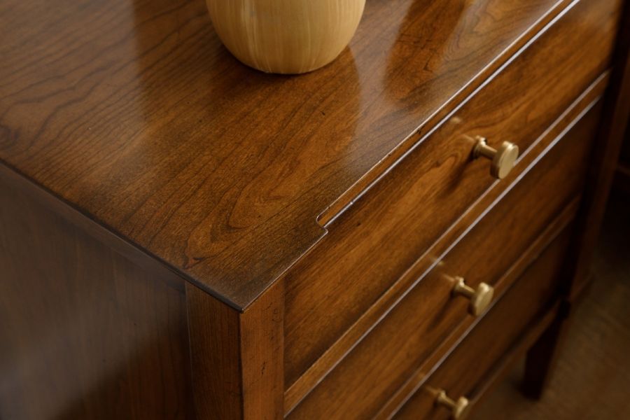 Close-up view of a wooden dresser surface showing the intricate wood grain patterns with a soft sheen, alongside brass drawer pulls and the edge of a turned wooden vase resting on top.