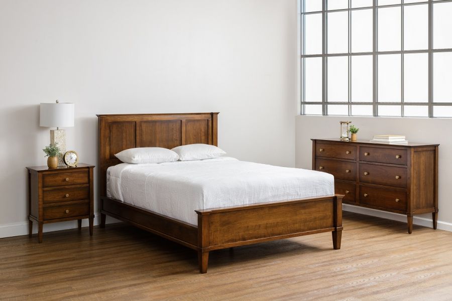 The Toulon Bedroom Set featuring a queen-sized bed with a wooden headboard, flanked by a matching nightstand and dresser, all in a rich wood finish, arranged in a bright room with a large window.